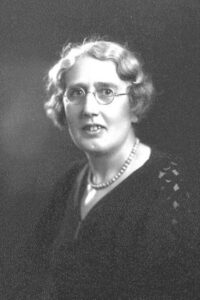 Black-and-white portrait of Dr Catherine Chisholm, wearing round glasses and a beaded necklace, with short wavy hair, facing slightly to the side.
