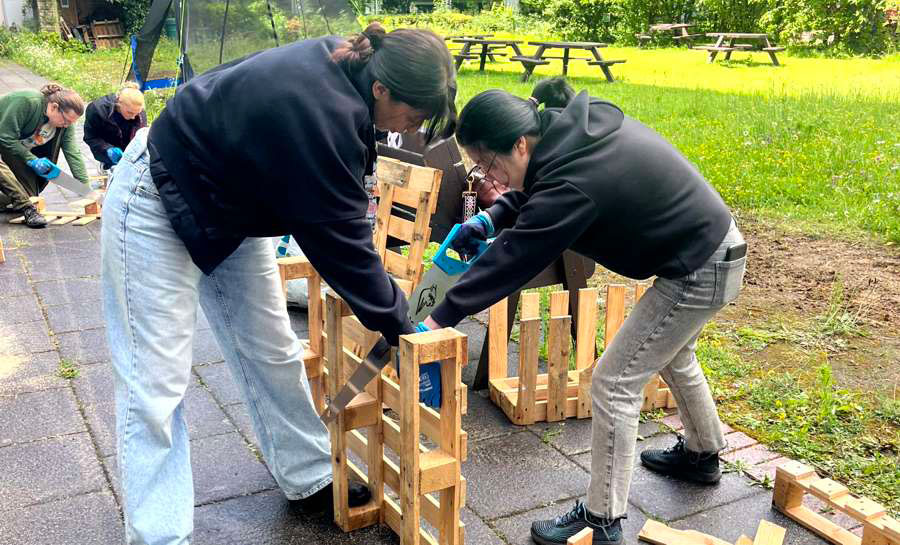 Two participants sawing a wooden pallet with a handsaw, with a half-finished flower planter beside them and the Michael Smith Quad in the background.