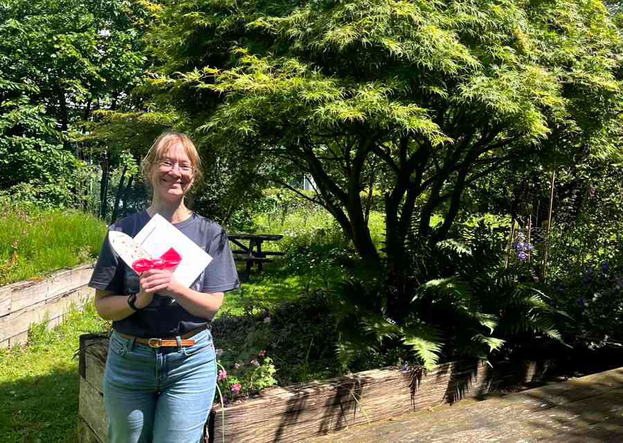 Emma in front of her raised bed in the Michael Smith Quad. [Alt text: Quad gardener winner, Emma Keevill, standing in front of her raised bed (number 3), holding a white trowel with a bee pattern and an envelope of FoodOnCampus vouchers.