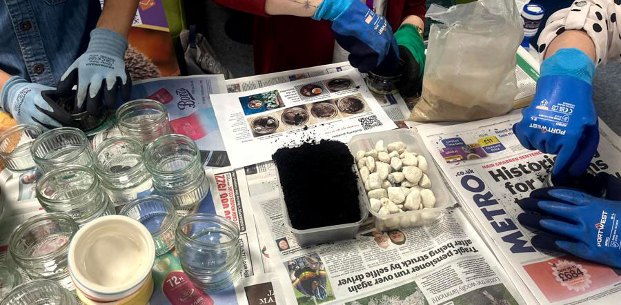 Glass ramekins and containers of compost pebbles and sand on a newspaper-lined table, with three pairs of gloved hands pressing compost into separate ramekins to create insect puddlers.