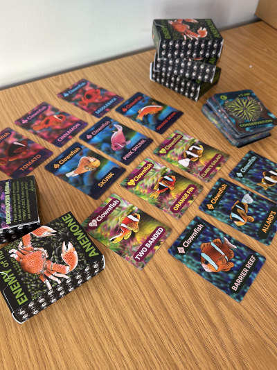 Enemy of My Anemone card game laid out on a wooden table, with colourful illustrated marine-life cards and several stacks of cards.