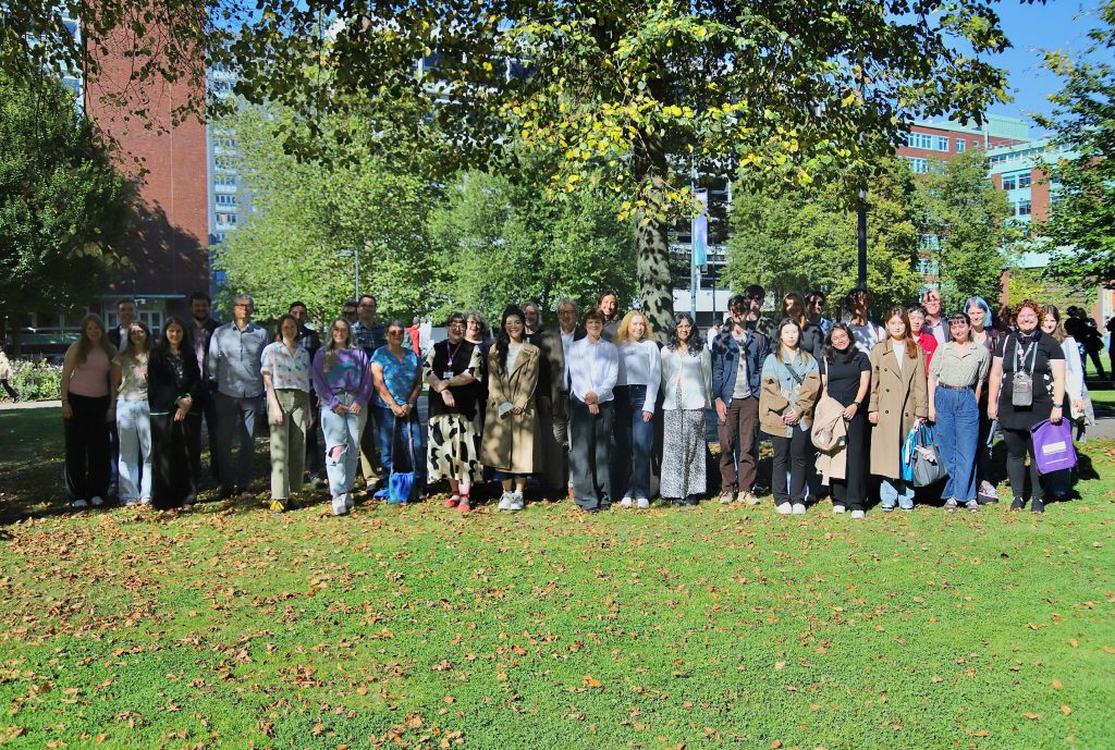 Photograph of a group of students and staff on a lawn under trees, with university builidings in the background