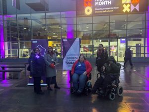 Rachel Heyes, Jackie Carter, and Laura Howard stand in front of the Alan Gilbert Learning Commons. Kathryn Bradley and Hamied Haroon use wheelchairs. Laura uses a crutch. All are smiling and are dressed in big coats with scarves and hats. It's dark, and so the purple light coming from the building illuminates the group.