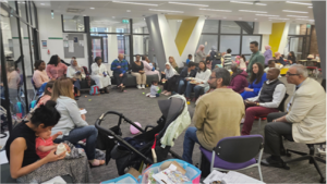 A group of adults (parents) some of the with small babies on their knees, are sitting in a circle. on chairs