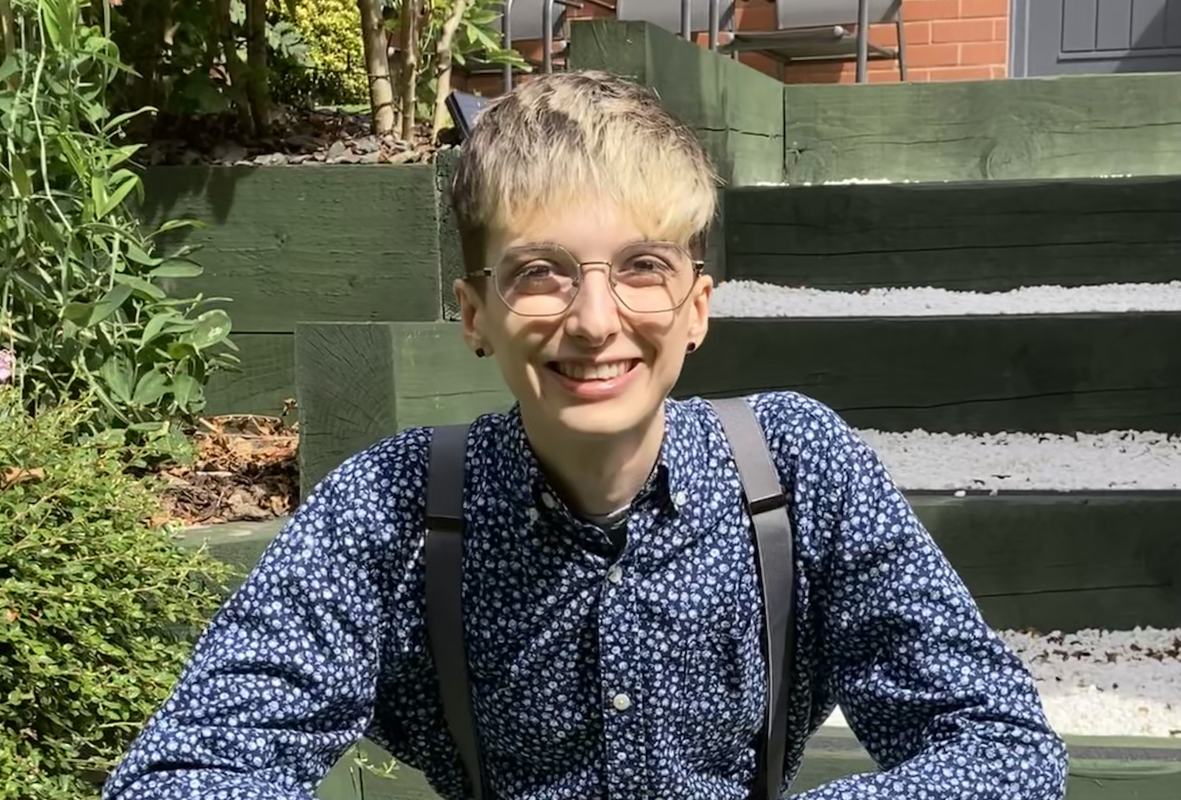 Beck Chamberlain Heslop with short blond hair wearing wire frame glasses smiling at the camera, in a blue shirt