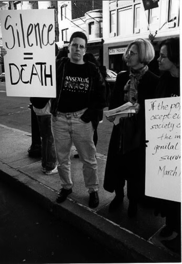 “Three people standing on a sidewalk during a protest. One holds a sign reading ‘Silence = Death,’ and another a partially visible sign in the background contains fragmented text.”