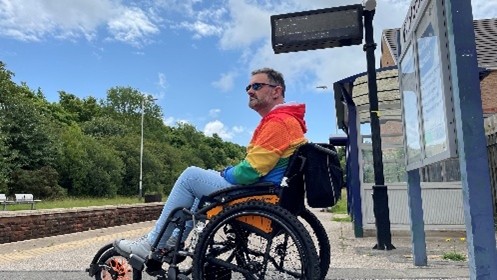 A person wearing a rainbow-coloured jacket and light blue jeans sits in a wheelchair at an outdoor train station platform. There is a digital sign overhead, a shelter to the right, and green trees in the background under a partly cloudy sky.