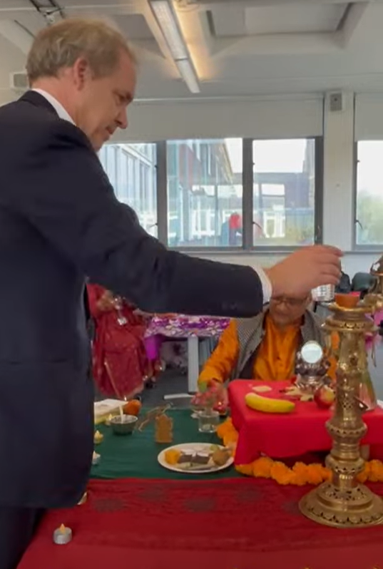 A person in a suit is lighting a traditional brass lamp on a decorated table during a cultural ceremony. The table is adorned with marigold flowers, fruits, and ritual items, while another individual in orange attire sits nearby. The setting appears to be indoors with large windows in the background.