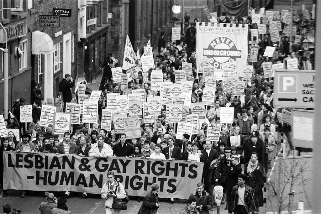 A large crowd marches down a city street in a black‑and‑white photograph, holding signs and banners supporting LGBTQ+ rights. At the front, a wide banner reads “LESBIAN & GAY RIGHTS = HUMAN RIGHTS.” Numerous placards behind it display slogans advocating equal rights and protesting discrimination. The crowd fills the street, with buildings and shop signs lining both sides.