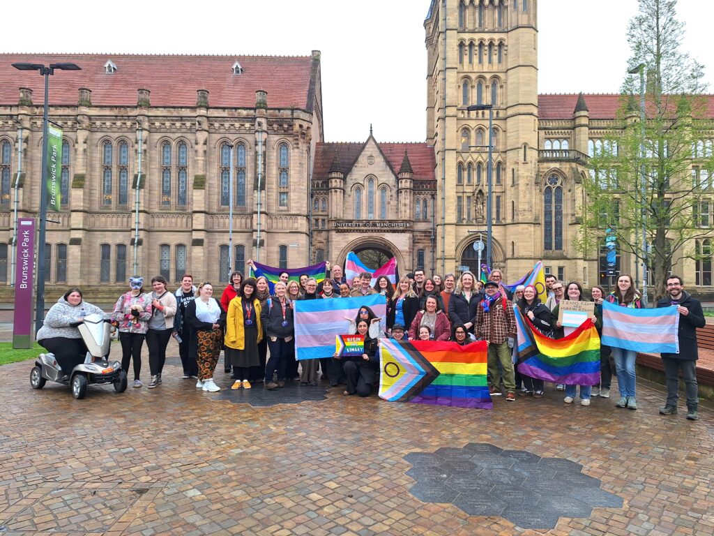 A large group of people standing outdoors in front of the University of Manchester’s historic main building, holding Pride, trans, and Progress Pride flags. The group is photographed on a paved square with the university archway and stone buildings in the background.