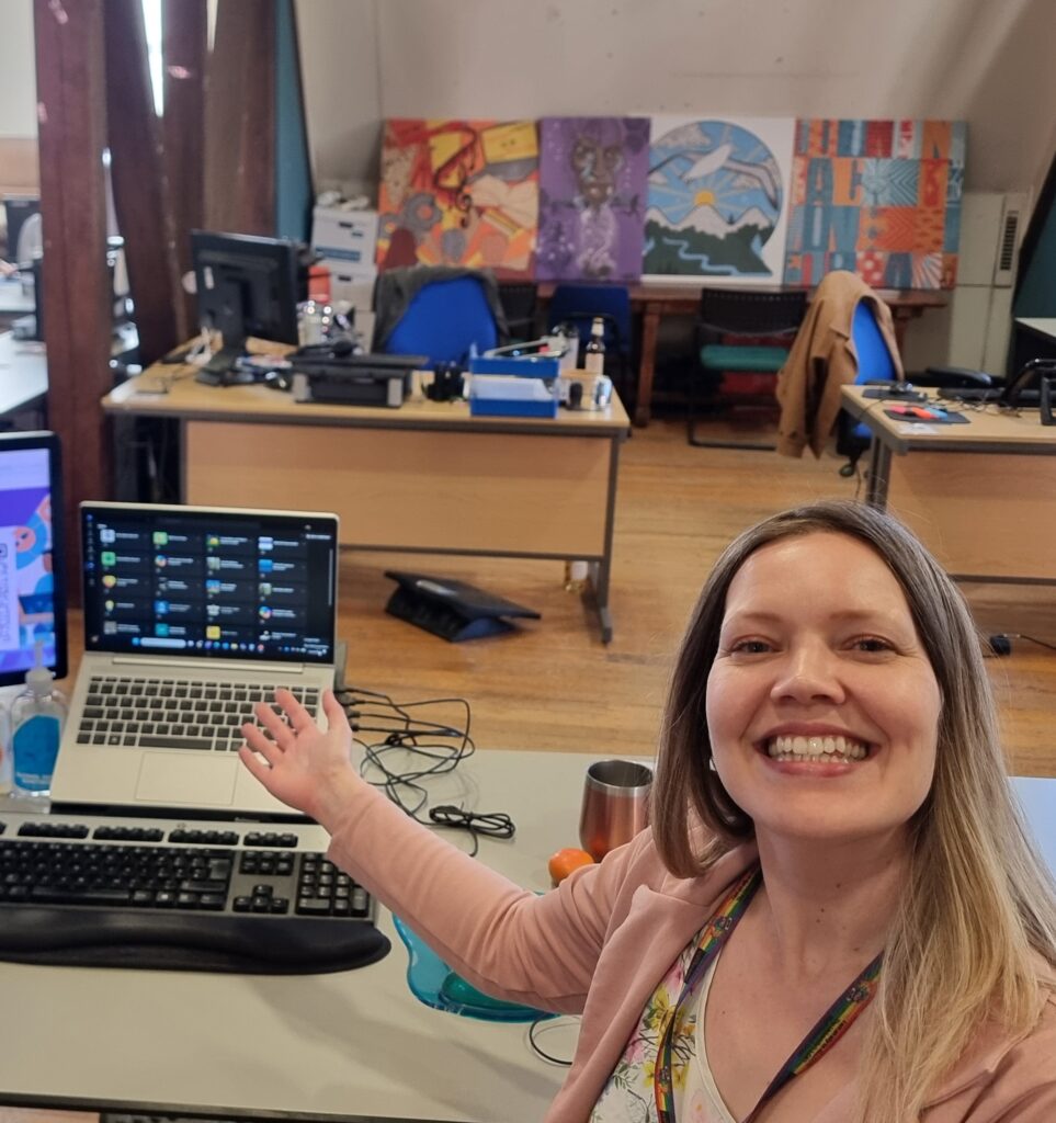 Rebecca Evans. Person standing behind a desk with arms outstretched in an open-plan office, photographing themselves. In the foreground is a workstation with a laptop and keyboard. Behind the desk are several workstations, blue office chairs, and large colourful artworks displayed along the back wall.