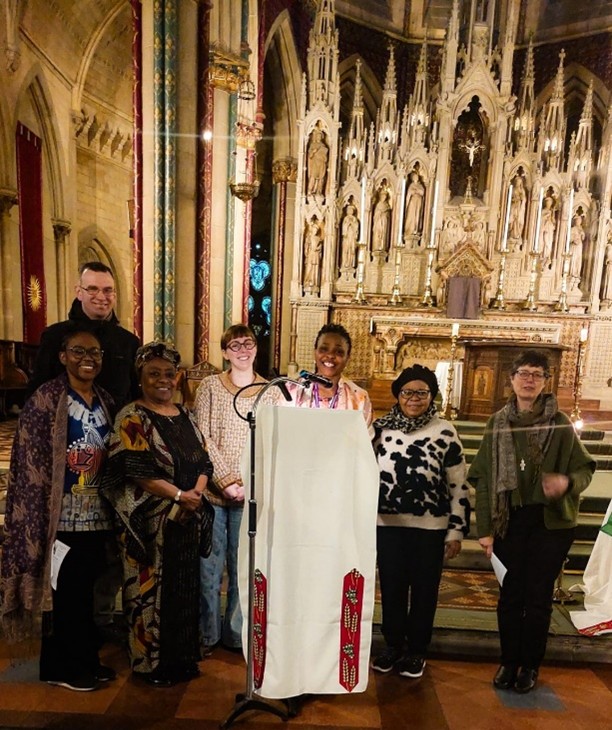 Group of people standing at a lectern inside a church or cathedral. The lectern with a microphone is at the centre, and the group stands in front of an ornate altar with carved figures and decorative panels. The interior features stone arches, patterned columns, and warm lighting.
