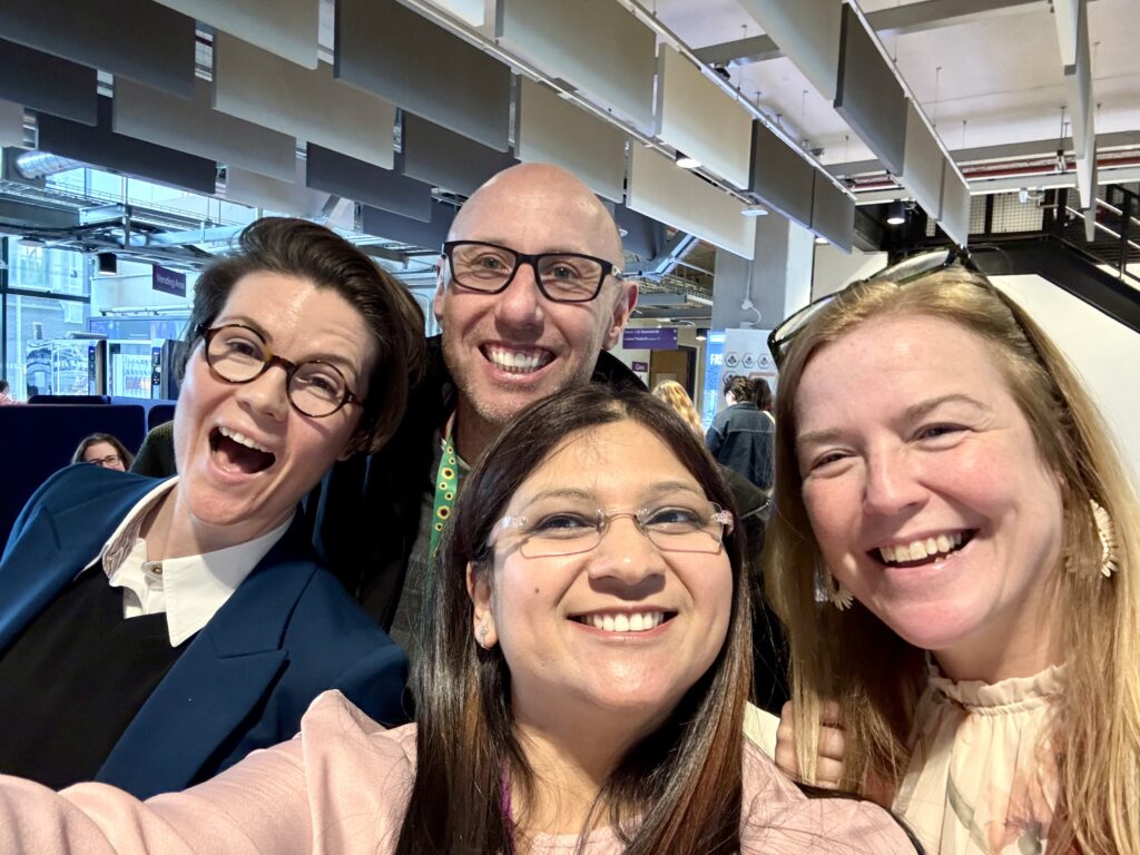 Four people take a close-up group selfie in a modern indoor public space with high ceilings and hanging acoustic panels. All four are positioned tightly together in the foreground, smiling toward the camera. One person at the front holds the camera, with three others leaning in from the sides and behind. Several of the individuals are wearing glasses. Behind them, the background shows a busy venue with signage, people walking, and architectural features such as exposed ducts, lighting fixtures, and suspended panels. The lighting is bright and even, suggesting a well-lit indoor environment.