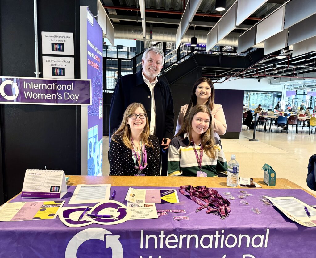Four people are gathered behind an information table at an indoor university venue during an International Women’s Day event. Two people are seated at the front of the table and two are standing behind them, all facing the camera. The table is covered with a purple cloth and displays leaflets, sign-up sheets, pins, lanyards, and promotional materials. A large sign to the left reads “International Women’s Day,” with additional smaller signs above for staff networks, including “Women@Manchester Staff Network” and “Endometriosis/PCOS Staff Network.” The background shows a modern, open space with high ceilings, exposed ducting, suspended acoustic panels, and people seated at tables further back.