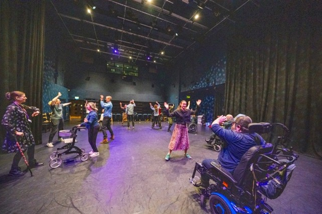 A group of participants, including wheelchair users, taking part in a dance session in a theatre space, with raised arms and movement across a large stage.