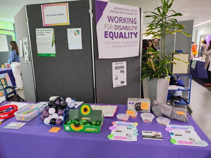 An information stall with a purple tablecloth displaying resources, sensory items, and leaflets about hidden disabilities, with posters behind including “All minds matter” and “Working for Disability Equality.”