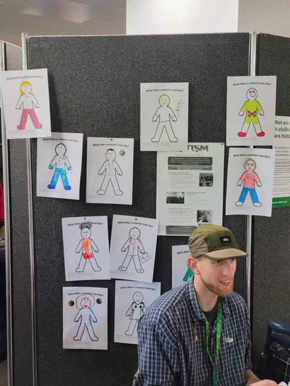 A display board showing multiple drawings responding to the prompt “What does a scientist look like?”, alongside informational posters, with a seated attendee in front of the display.