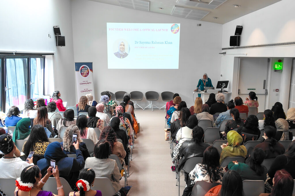 Wide view of a conference room during a presentation. An attendee stands at a lectern addressing a seated audience, with a presentation slide and event banner visible at the front of the room.