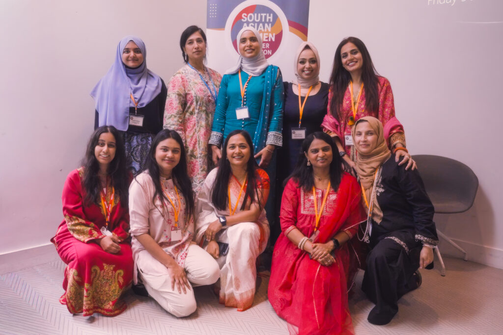 Group photograph of ten event participants standing and kneeling in front of a South Asian Women in Higher Education banner, wearing conference badges and traditional and contemporary clothing.