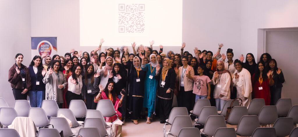 Large group photograph of event attendees gathered at the front of a conference room. Participants are standing together and waving towards the camera beneath a projected screen.