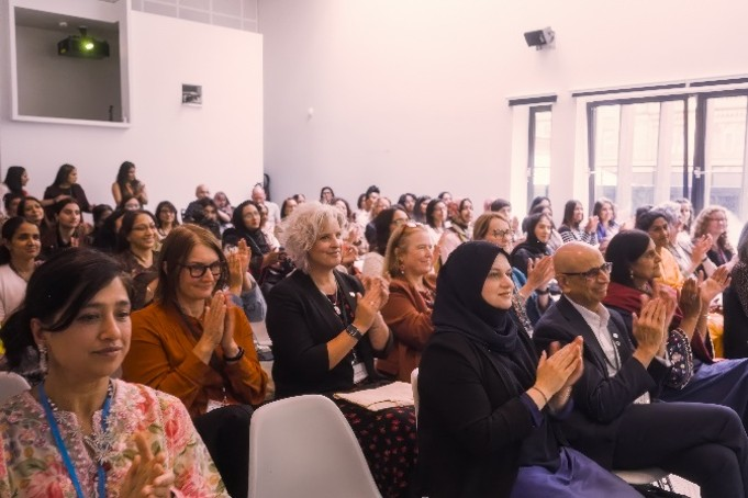 Audience members seated in a conference room applauding during a South Asian Women in Higher Education event. Attendees are arranged in rows facing the front of the room, with large windows allowing natural light into the space.
