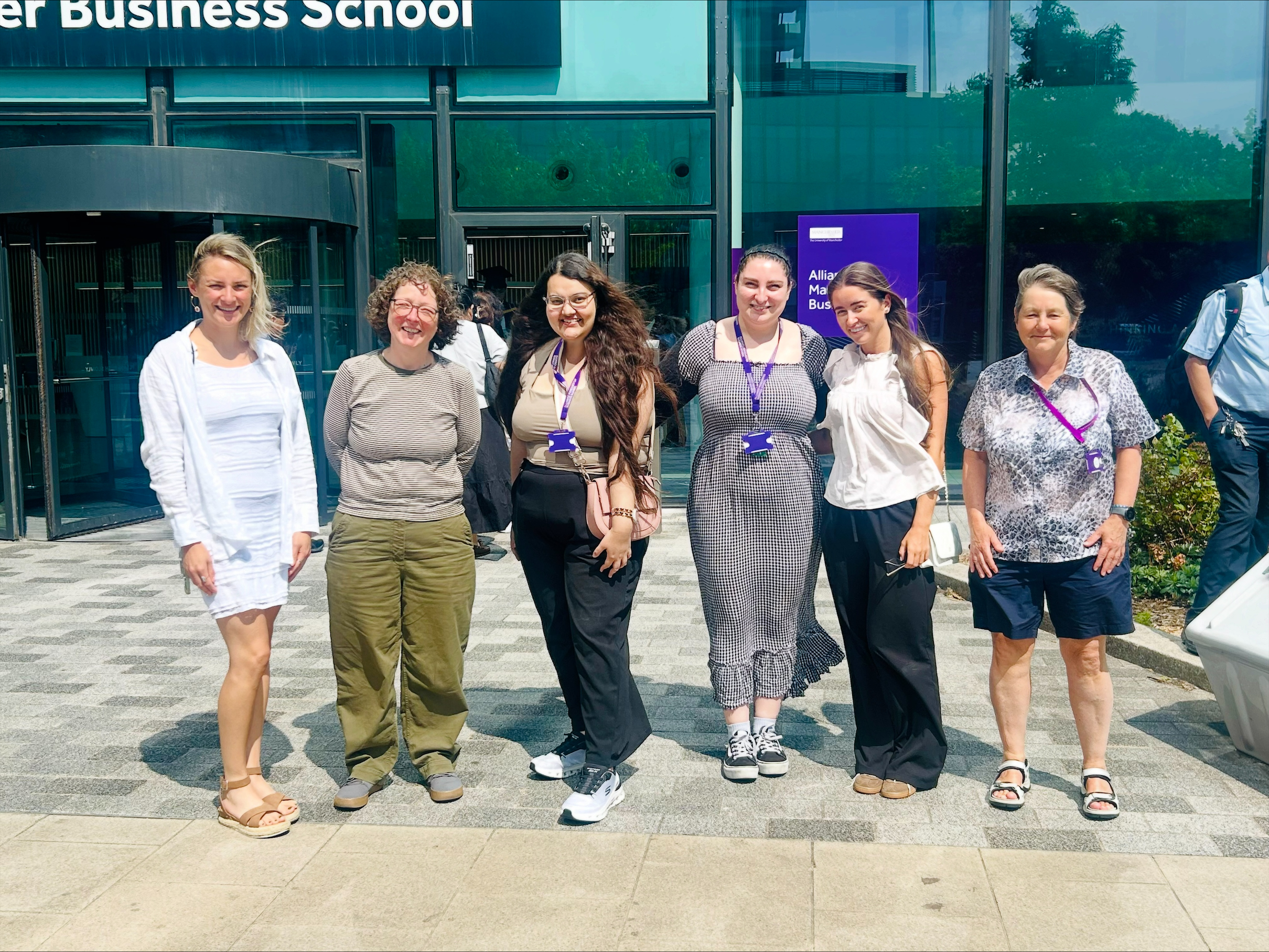Group photo of six people standing outside the entrance of a business school building on a sunny day. The group is posed in front of glass doors and windows, with signage for the business school visible in the background.