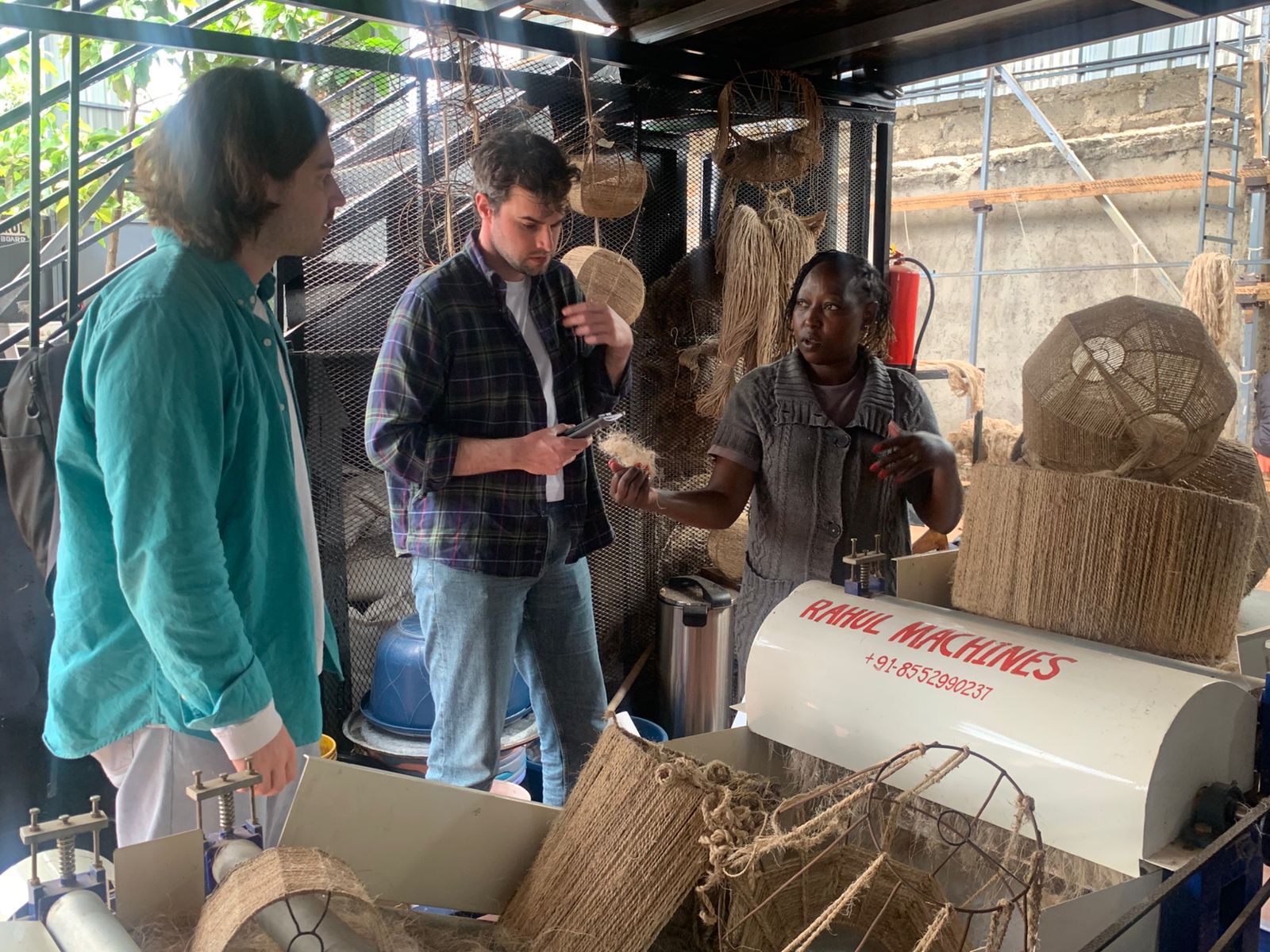 Three people stand around a fibre-processing machine in a workshop, with woven baskets, natural fibres and industrial equipment visible around them.