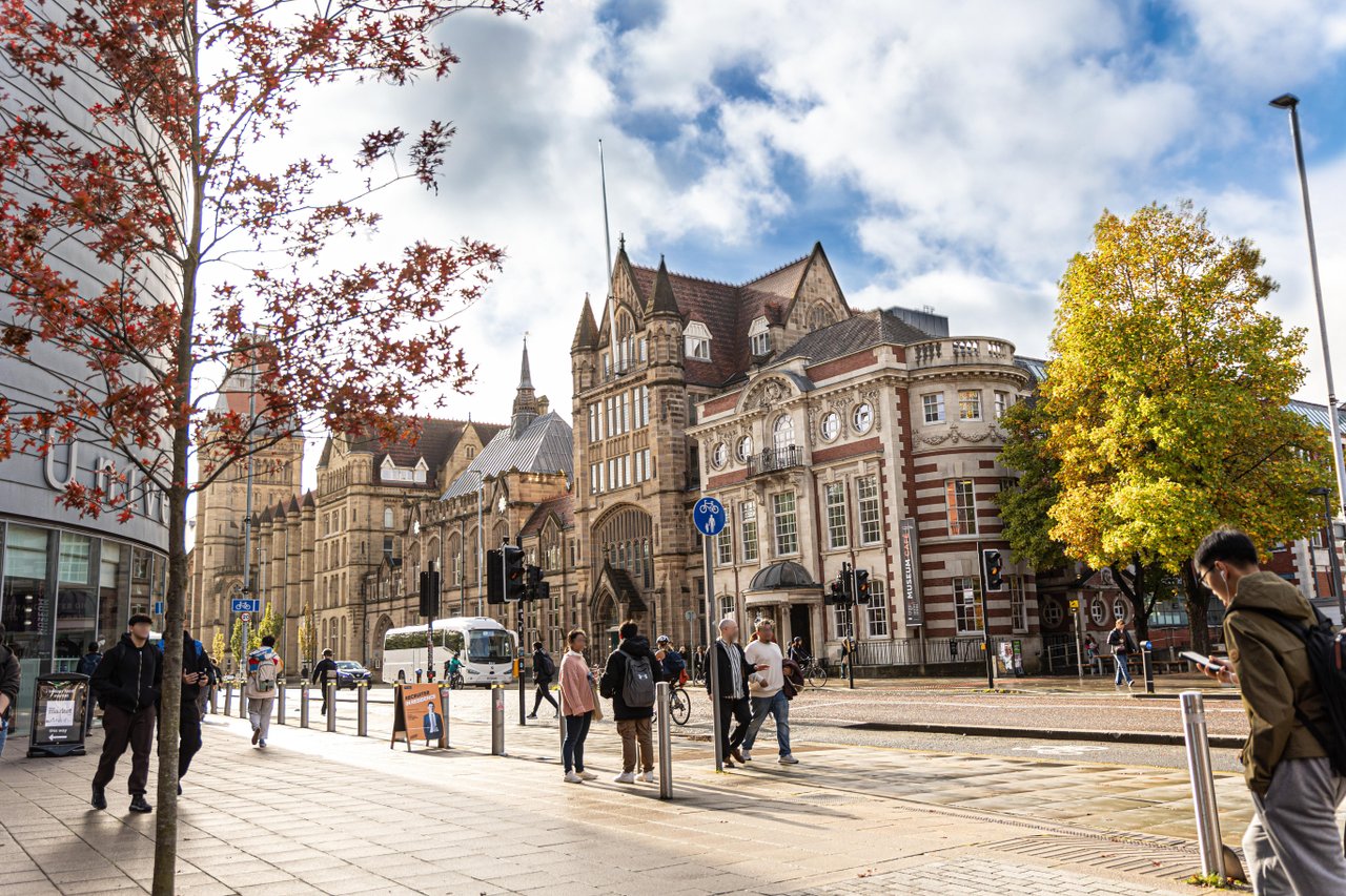 A busy city street lined with ornate historic stone and red-brick buildings, with pedestrians standing and walking along the pavement and crossing the road, a bus and bicycles nearby, and autumn trees beneath a partly cloudy blue sky.