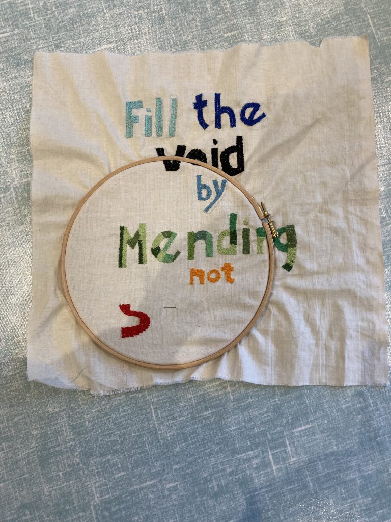 Embroidered fabric in a hoop with the message "Fill the void by mending, not spending," promoting repair and sustainable fashion practices.