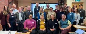 Group of people smiling and posing indoors during a workshop or meeting, with name tags and laptops visible, in front of a screen displaying 'JUST'