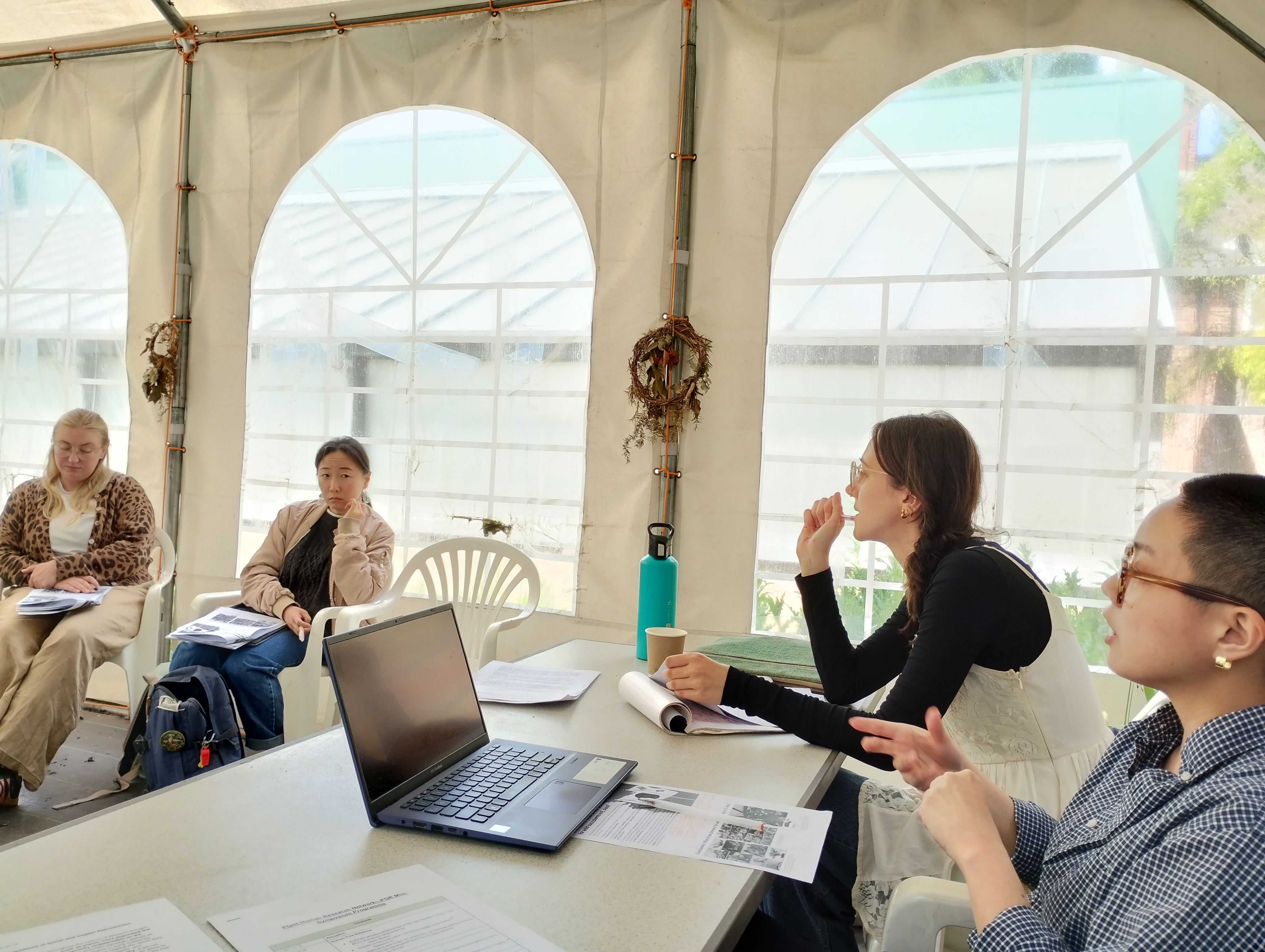 Four people engaged in discussion inside a garden marquee, seated around a table with laptops, printouts, and natural wreaths hanging on the tent walls.