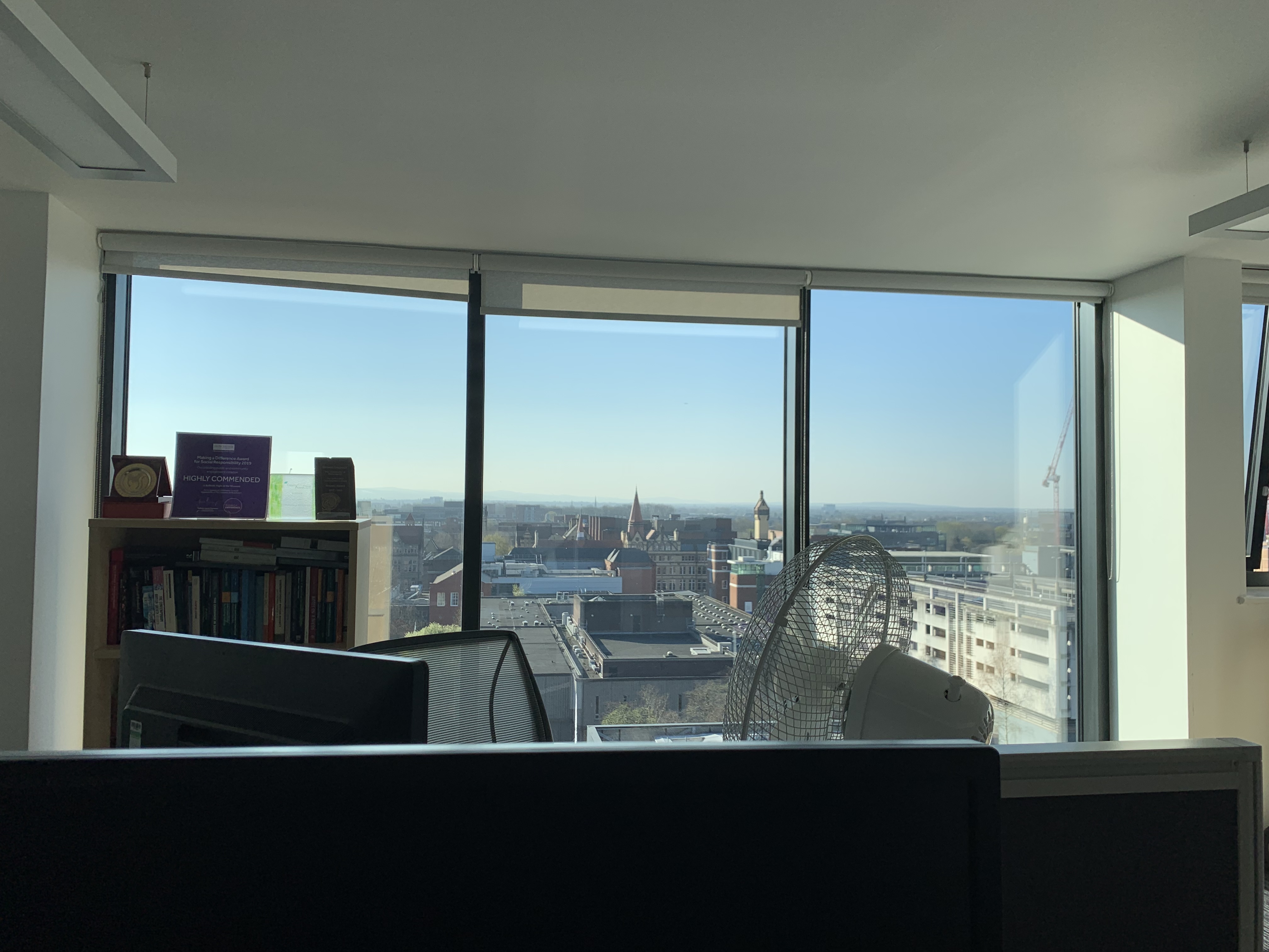 Office with bookshelf, fan, and computer monitors in front of large windows overlooking Manchester cityscape on a clear, sunny day.