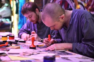 Two individuals stamping and working on craft projects at a table covered with tools and paper.