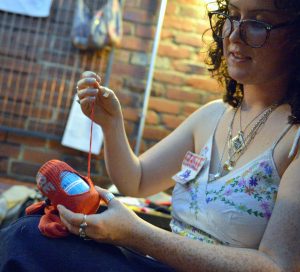 Person threading a needle with red yarn, holding a ball of yarn in a cozy brick-walled setting.