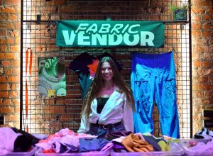 Fabric vendor booth with clothes and textiles displayed against a brick wall under a green banner.