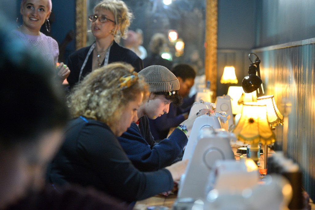 Group seated at a long table using sewing machines under warm lamp light in a workshop.