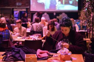 Individual hand-stitching fabric at a table during a communal crafting event with others in the background.
