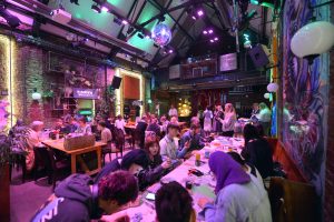 Large indoor venue with colorful lighting hosting a sustainable fashion workshop; people seated at tables working on repair and upcycling projects