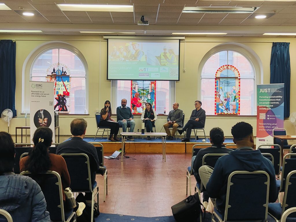 Panel discussion in a hall with five speakers on stage, audience seated, banners and stained glass windows in the background.