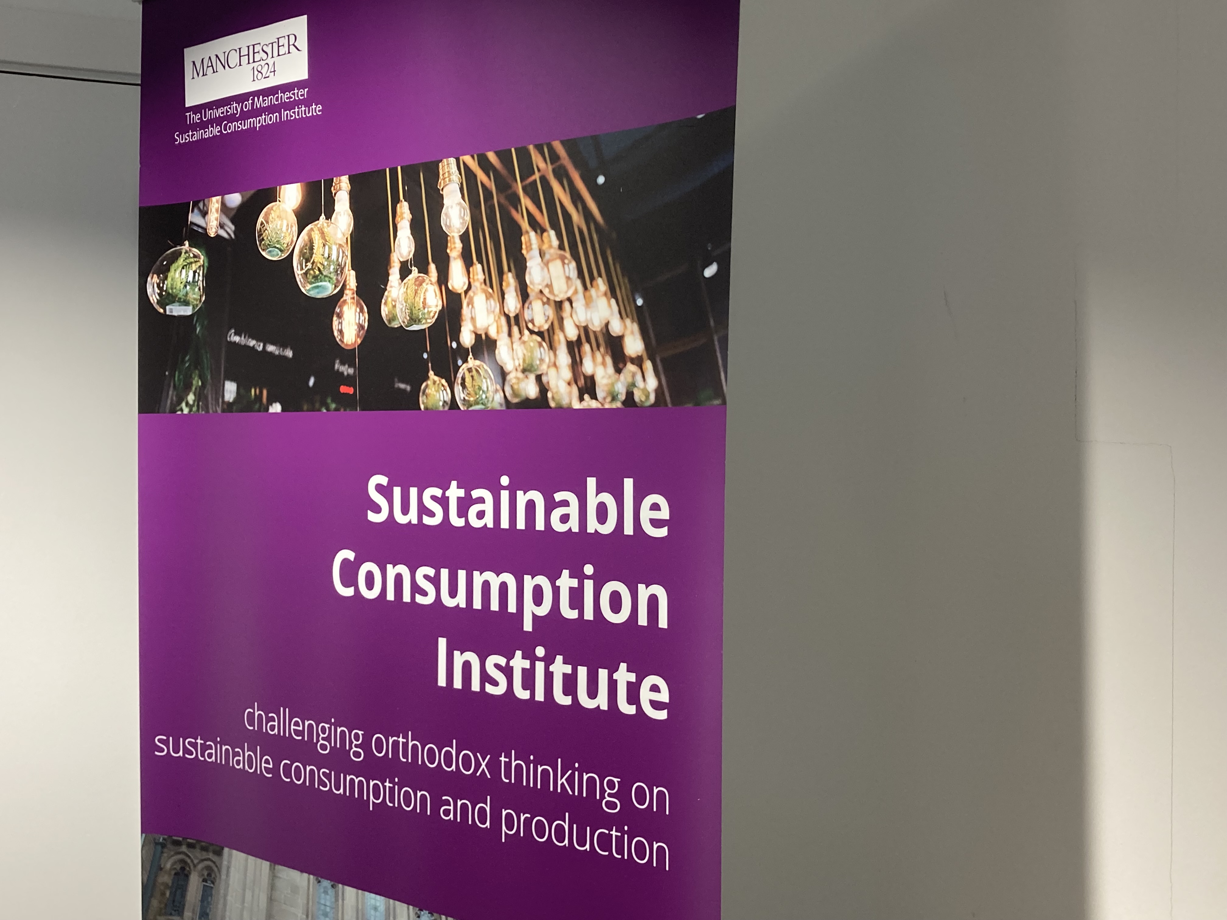 A purple banner for The University of Manchester’s Sustainable Consumption Institute, featuring hanging light bulbs with small plants inside and the tagline “challenging orthodox thinking on sustainable consumption and production.”